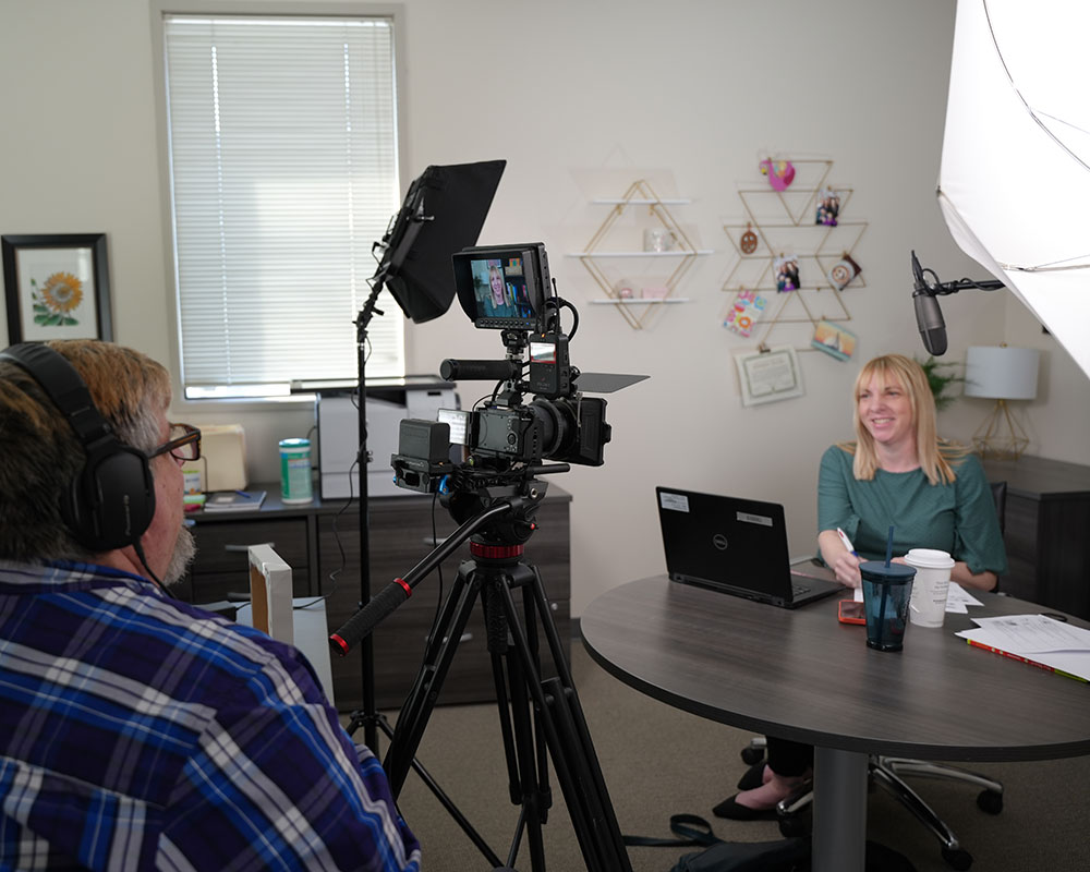 Molly Daley sits at a table and smiles for a videocamera.